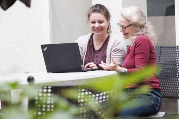 Zwei Frauen sitzen lächelnd vor einem Laptop am Tisch in einem Innenhof.